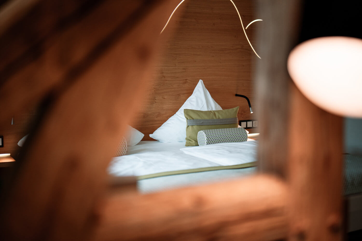 Blick durch rustikale Holzbalken auf ein gemütlich beleuchtetes Zimmer im Hotel Diedrich, Wellnesshotel Sauerland, mit stilvoller Bettwäsche und moderner Innenraumbeleuchtung, einladend für eine entspannte Übernachtung im Spa Hotel NRW.