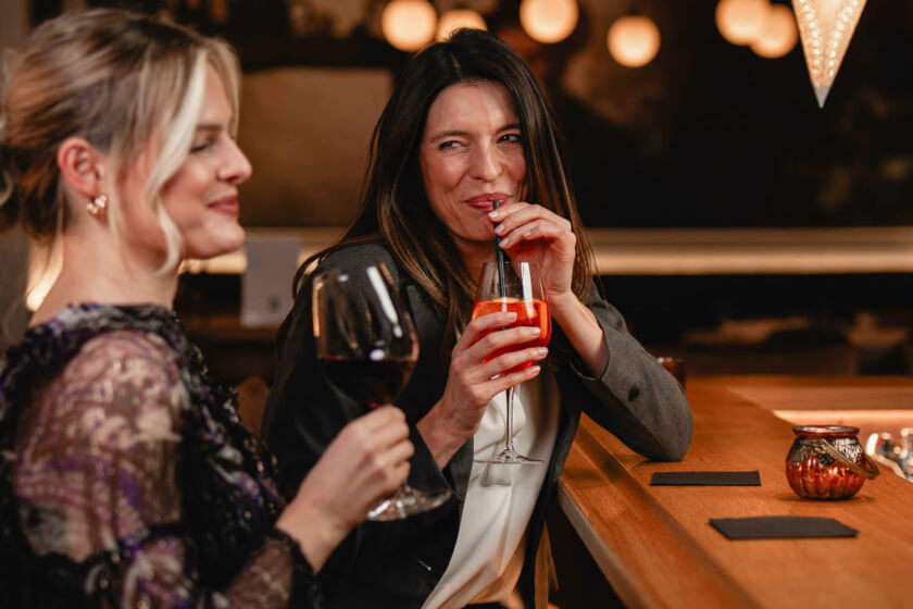 Eine Frau lächelt, während sie ein Getränk genießt, und unterhält sich mit einer Freundin an einer modernen Bar.