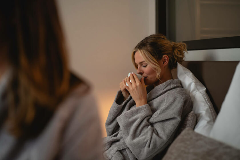 Eine Frau im Bademantel sitzt auf ihrem Bett und genießt den Duft ihrer Tasse Tee, die sie sich mit beiden Händen an den Mund hält.