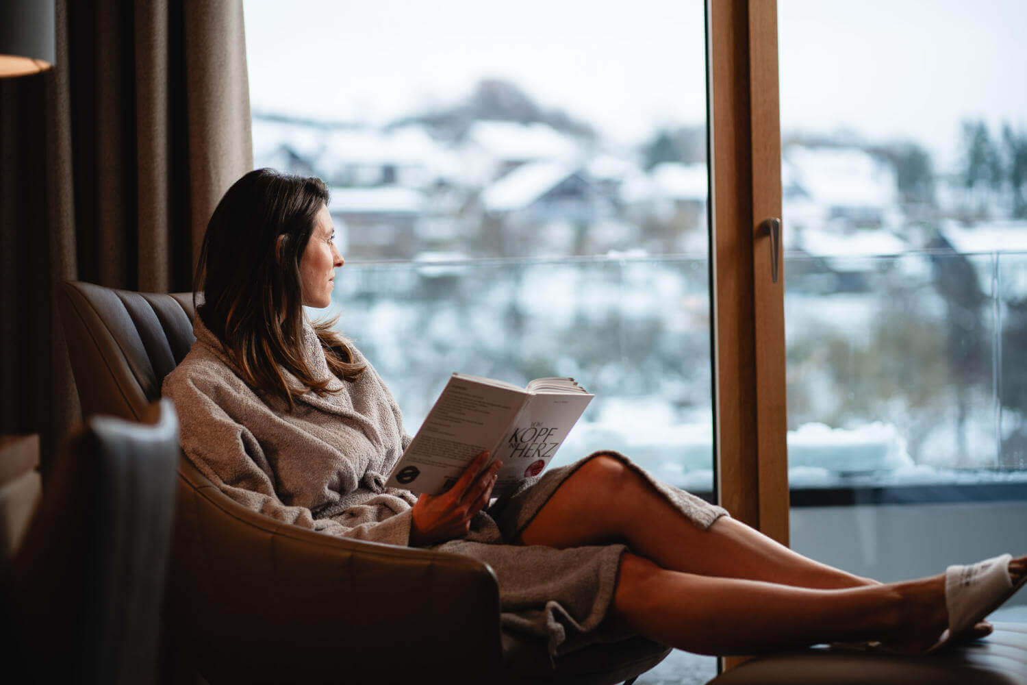 Eine Frau im Bademantel sitzt im Hotelzimmer auf dem Lesesessel und hält ein Buch in den Händen.